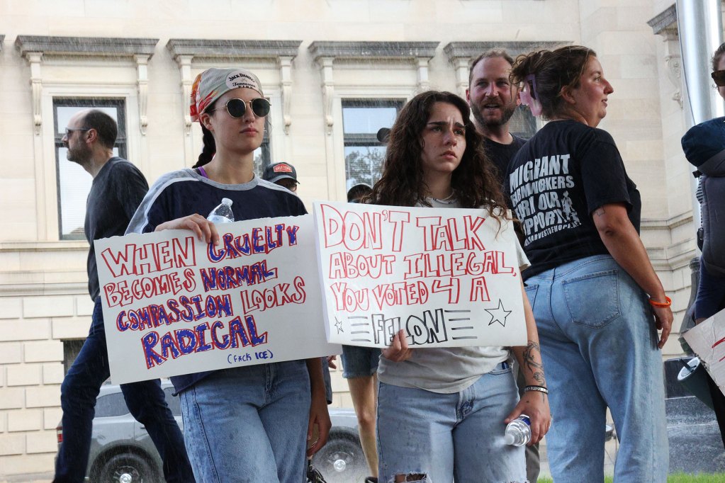 Protesters stand outside in the rain holding signs, one reads "When cruelty becomes normal, compassion looks radical"