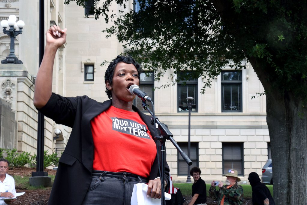 A woman in a shirt that reads "Your vote matters" speaks at a mic outside
