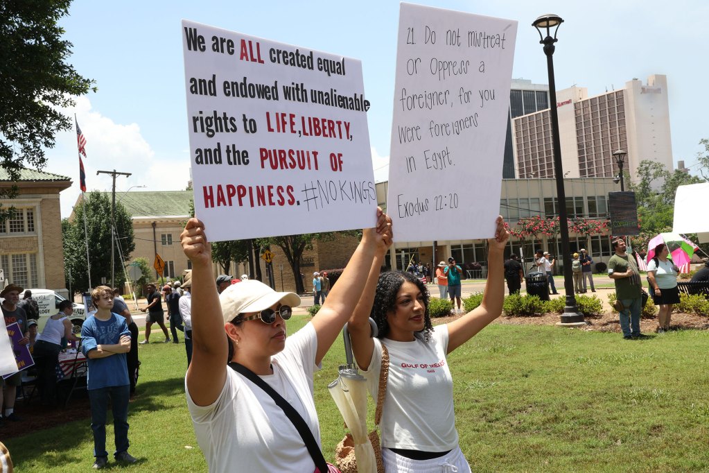 Protesters hold signs, one reads "We are all created equal and endowed with unalienable rights to Life, Liberty, and the Pursuit of Happiness. #nokings"