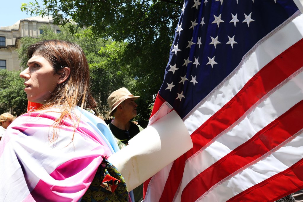 A woman stands outside draped in a transgender flag, US flag hangs behind her