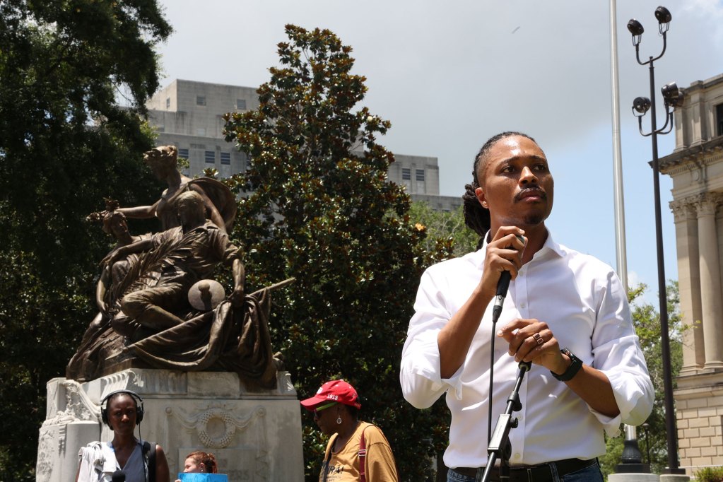 A man in rolled up sleeves talks at a mic outside, a large statue and trees visible behind him