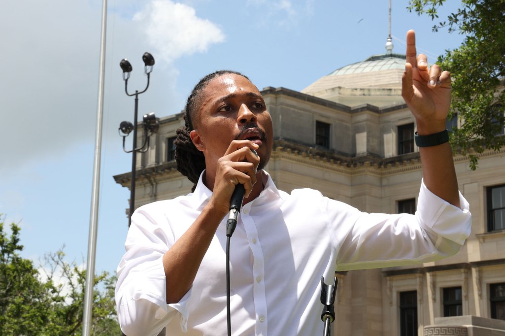 A man in rolled up sleeves talks at a mic outside, a large statue and trees visible behind him