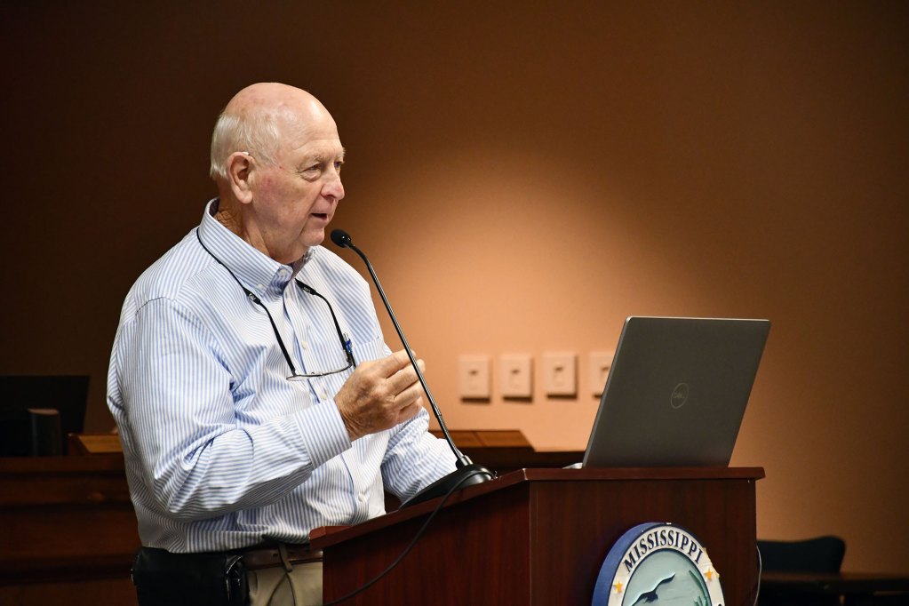 Joe Spraggins speaks at a podium with a laptop and microphone during an MDMR public meeting.