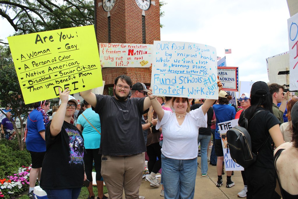 Protesters hold up signs, one reads "Our food is grown by migrant hands. While we rank last in learning plans. We deserve better MS. Fund Schools. Protect Workers. #NoKings"