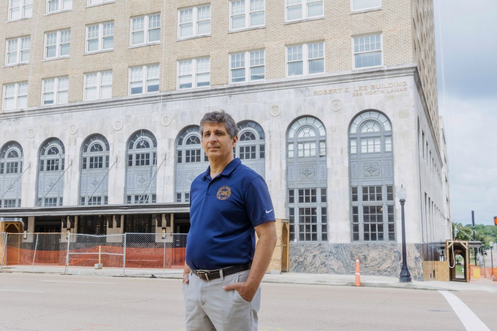 In a photograph, a White man with gray hair stands on the street in front of a concrete building with arched windows. The man is wearing a blue short-sleeved polo shirt with a logo.