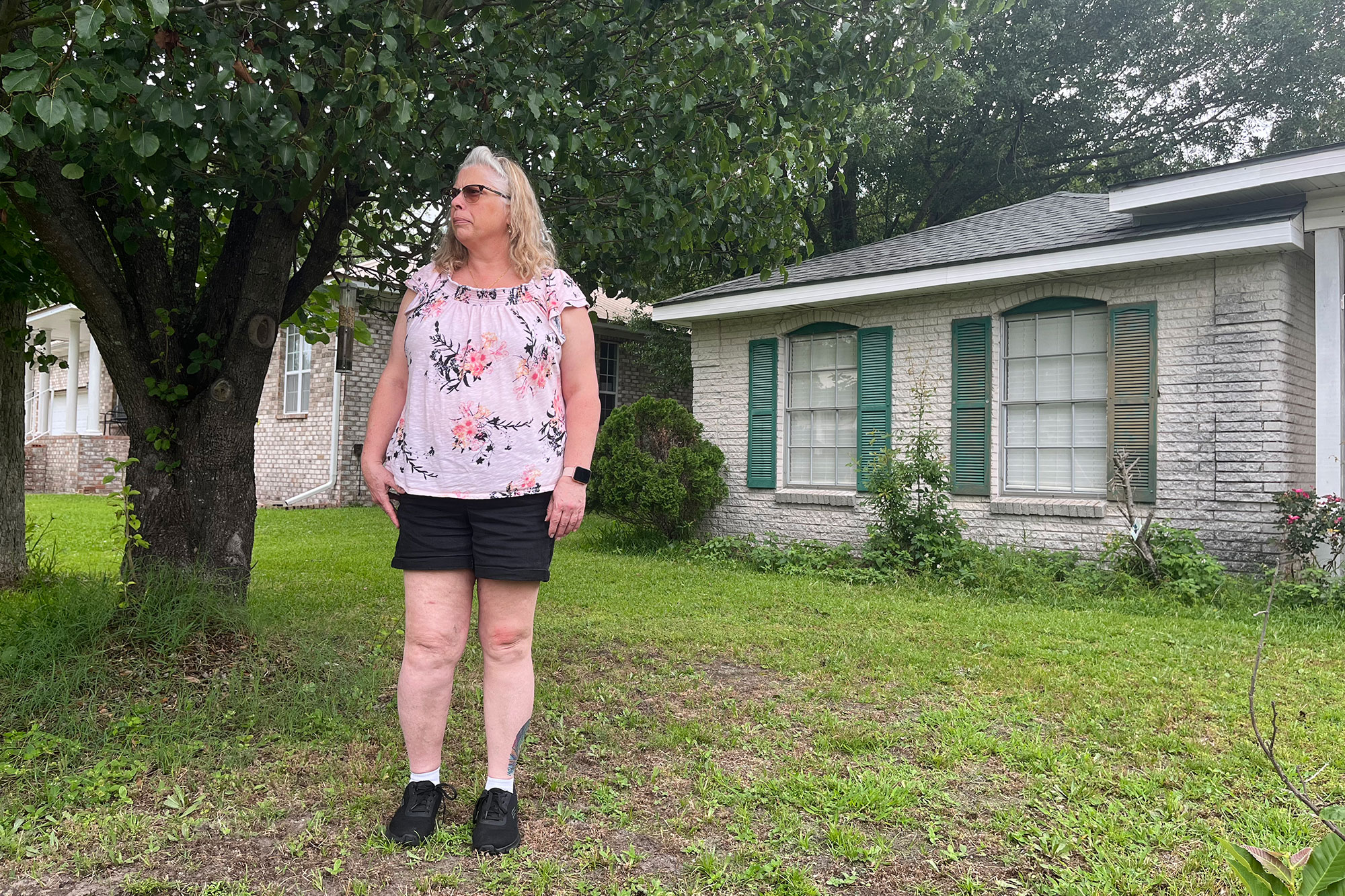 A woman stands in the yard of a white house with green shutters