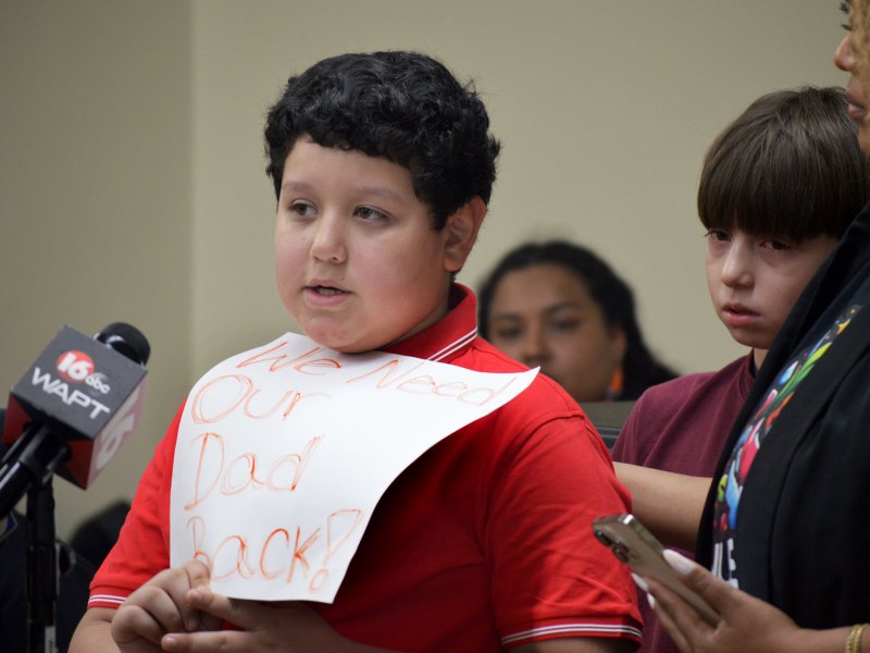Two young boys speak at a mic, one holding a hand written sign that reads "We Need Our Dad Back!"