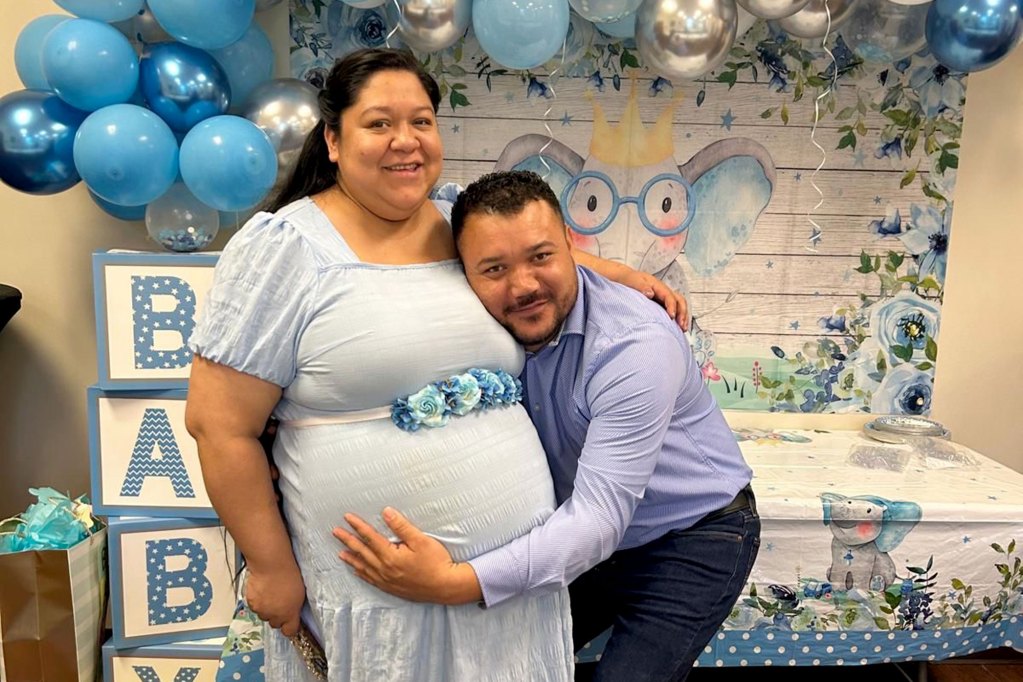 A man hugs the belly of a woman. Decorations for a baby shower are seen behind them.