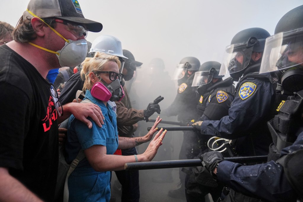 Protestors line up on the left against police lined up on the right. Everyone is wearing masks against the tear gas in the air.