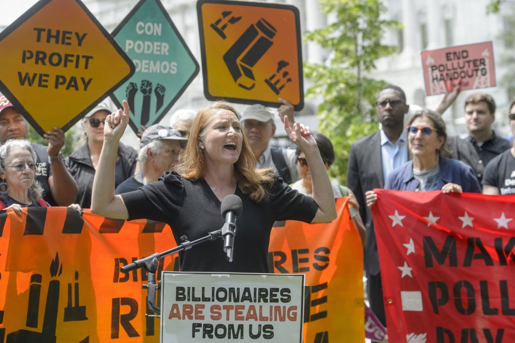 Melanie Stansbury speaks at a conference, signs can be seen that say "They profit we pay" and "Billionaires are stealing from us"