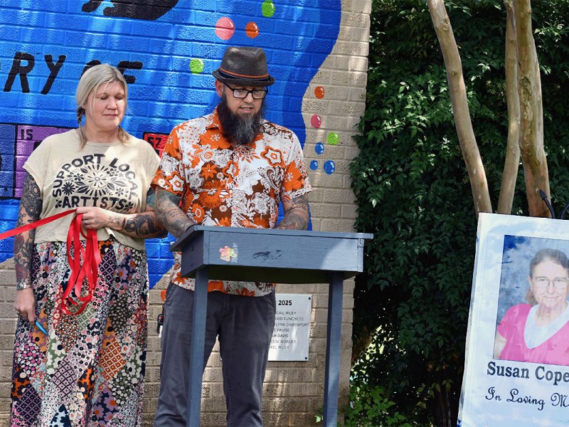 A man in a colorful shirt speaks in front of a mural. A photo of Susan Copeland is to the side, it reads In Loving Memory under her photo