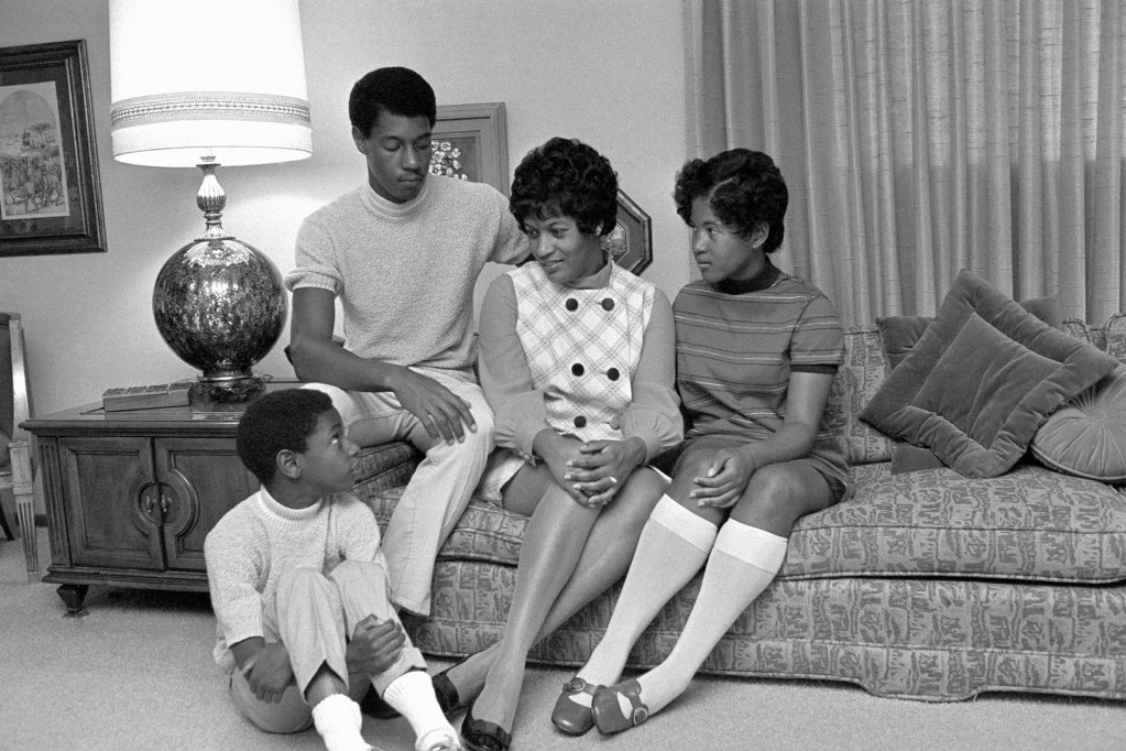 A black and white photo showing Myrlie Evers sits on the couch with her three children
