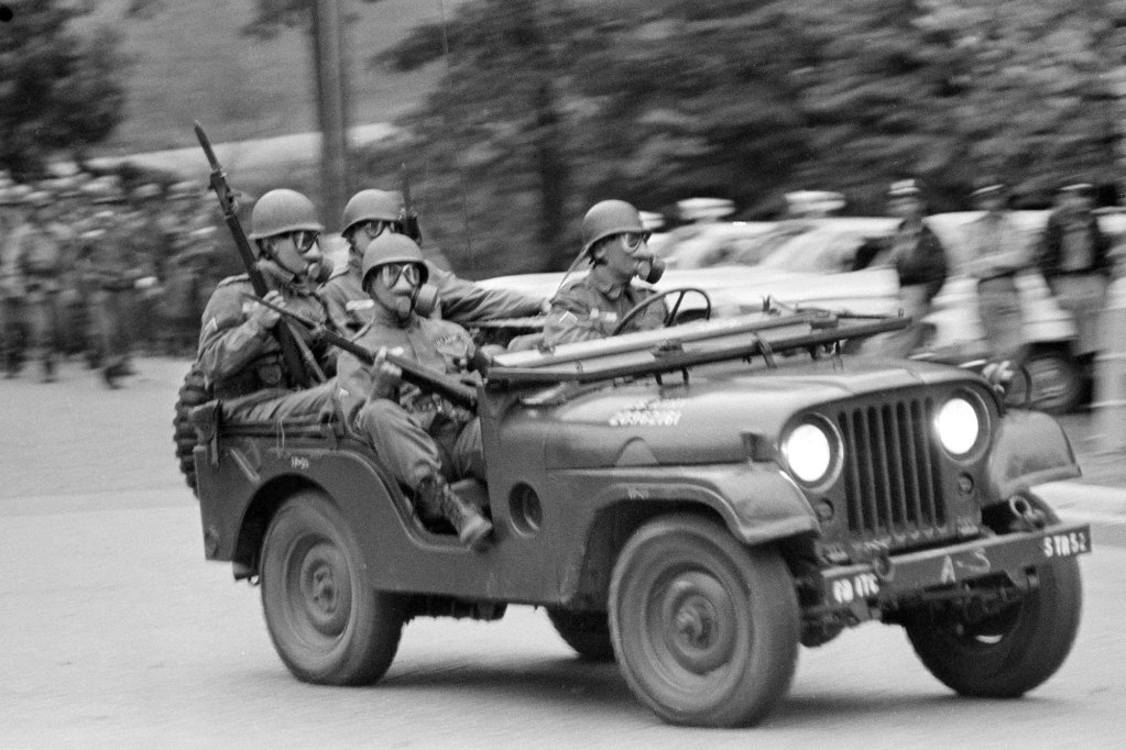 A black and white photo of a military jeep filled with four masked and armed soldiers driving by