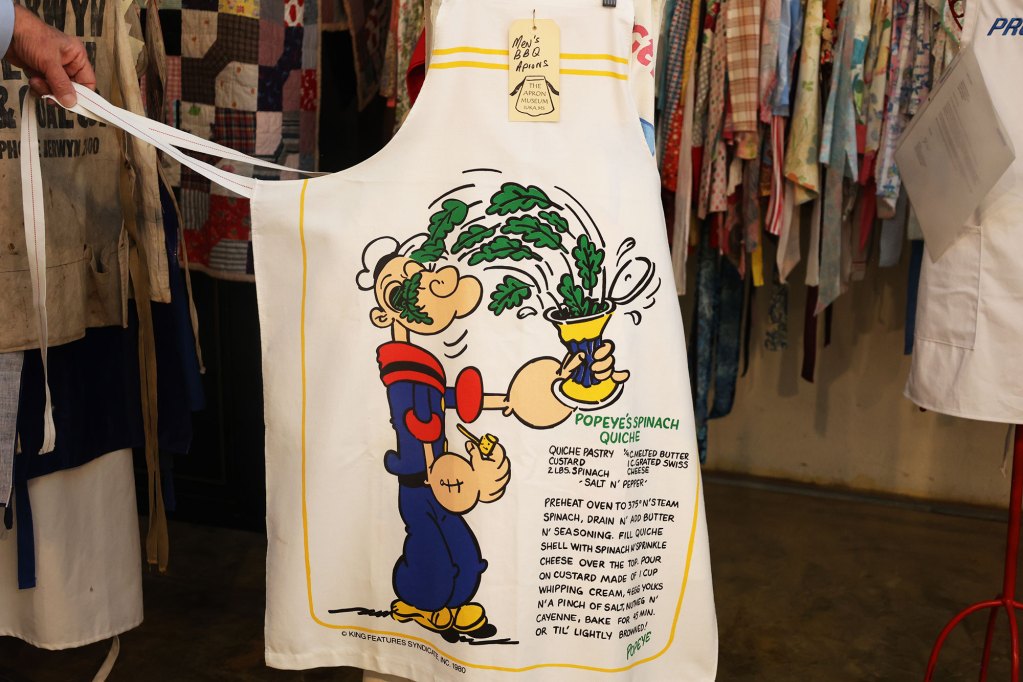 A white apron featuring the cartoon character Popeye beside a recipe for Popeye's Spinach Quiche