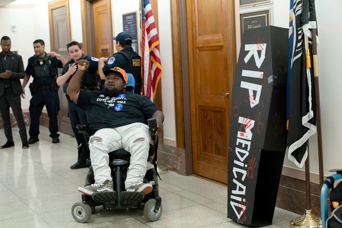 A demonstrator on wheelchair protests outside of a senator's office. A black coffin with white lettering that reads "RIP Medicaid" sits against the wall.