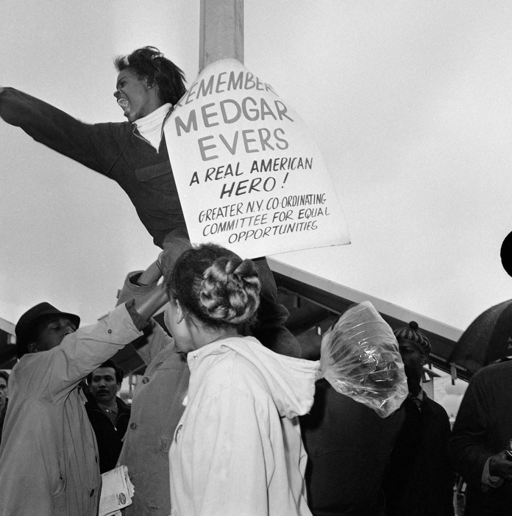 Civil rights demonstrator clings to pole outside holding a sign reading “Remembering Medgar Evers”
