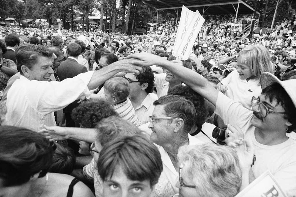 An old black and white photo from the 1980's of Ronald Reagan walking through a crowded political campaign event and shaking hands