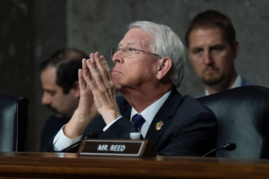 a photo of a white haired man listening with his hands clapsed in a committee room