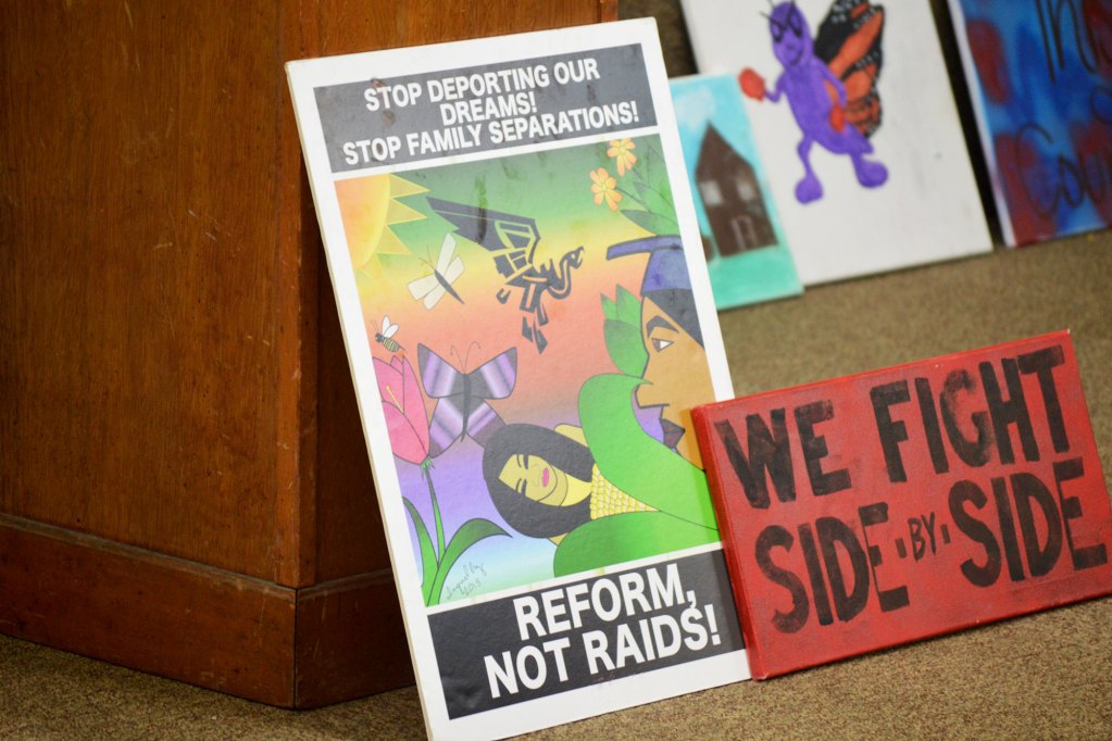 Signs lean against a podium that read "We fight side by side" and "Stop deporting our dreams! Stop family separations! Reform, not raids!"