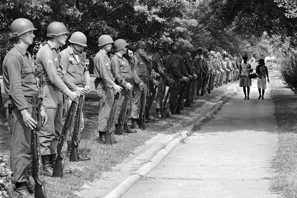 Uniformed soldiers sand in a line in the grass as two Black girls walk down the sidewalk
