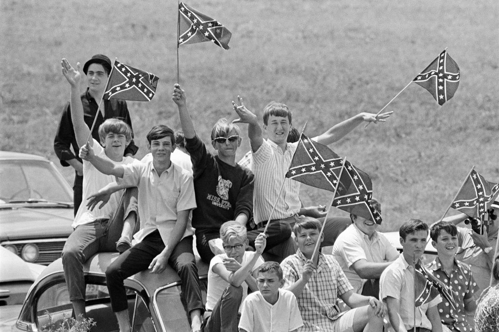 A white and black photo of white boys sit on top of cars waving small Confederate flags
