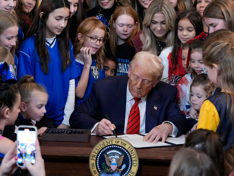 President Trump is signing a paper on his desk as many young girls look on