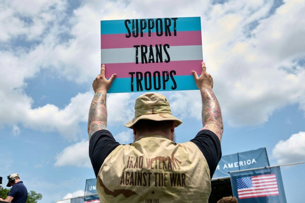A person wearing a vest identifying him as a veteran holds a sign up that says 'SUPPORT TRANS TROOPS.'