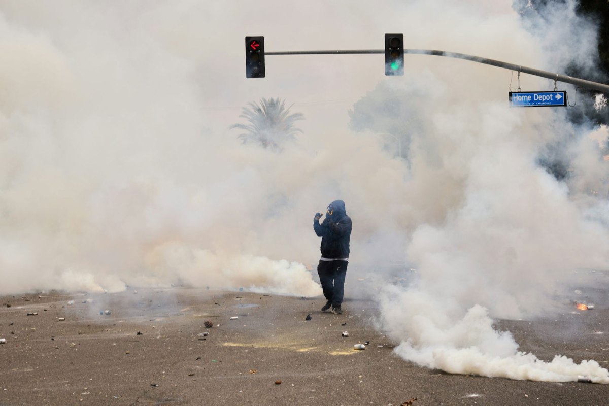 A single person in gas mask walks through a smoke filled road, the street sign reads Home Depot
