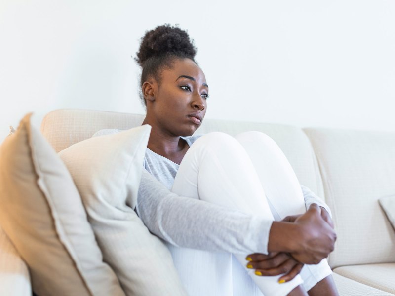 A sad woman dressed in white clothes seated curled up on a white couch