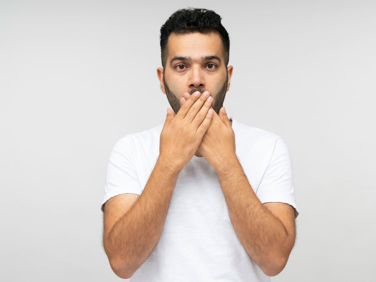 A man in a white top stands against a white background with both hands covering his mouth