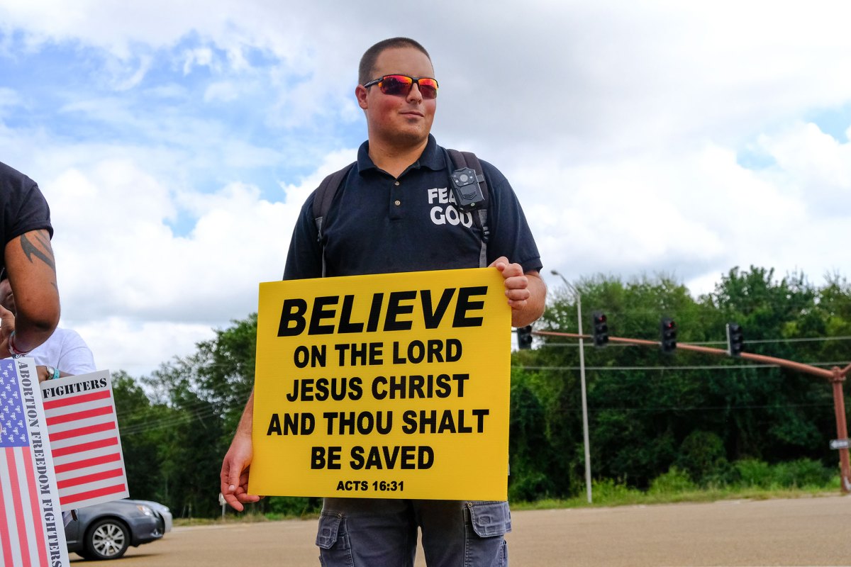 A protester holding a yellow sign that reads "BELIEVE ON THE LORD JESUS CHRIST AND THOU SHALT BE SAVED"