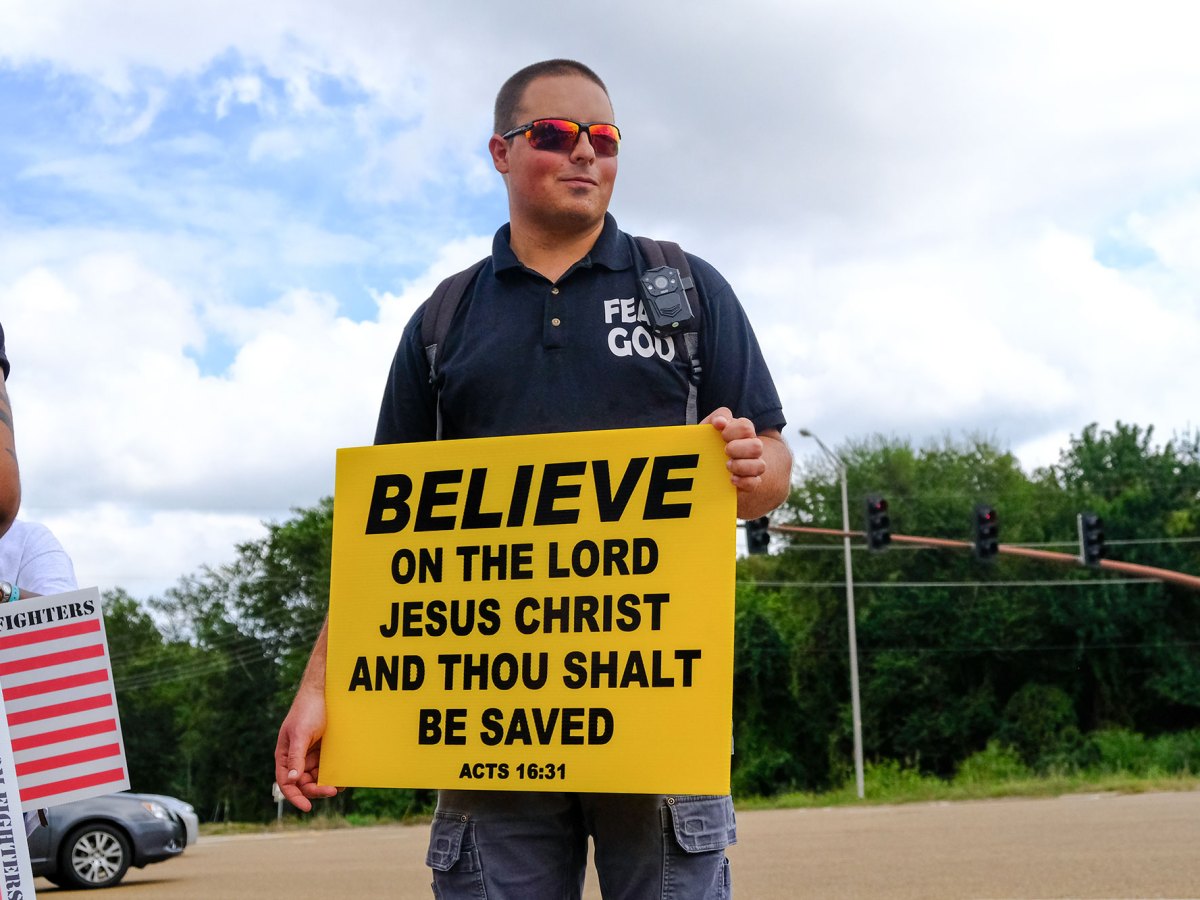 A protester holding a yellow sign that reads "BELIEVE ON THE LORD JESUS CHRIST AND THOU SHALT BE SAVED"