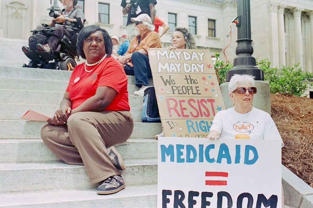 People sit on the stairs outside holding signs that read "May Day May Day"" and "Medicaid = Freedom"