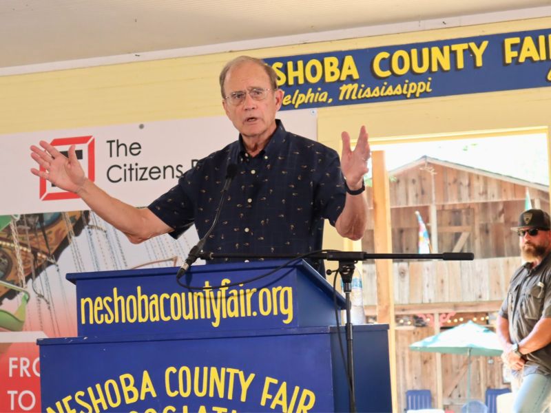 Delbert Hosemann speaks at a blue podium, arms held out wide. The podium reads "Neshoba County Fair" in yellow letters