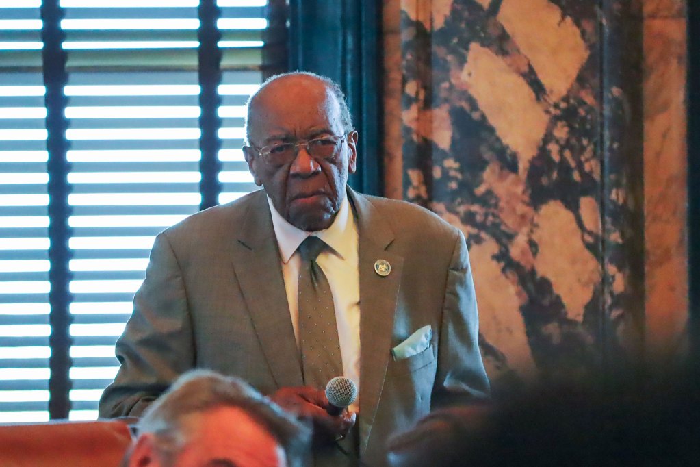 A man in a suit stands up with mic in hand. The walls behind him are marbled