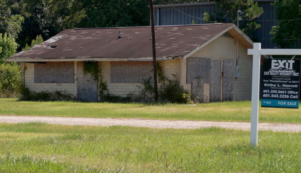 A for sale sign in front of a boarded up dilapidated house
