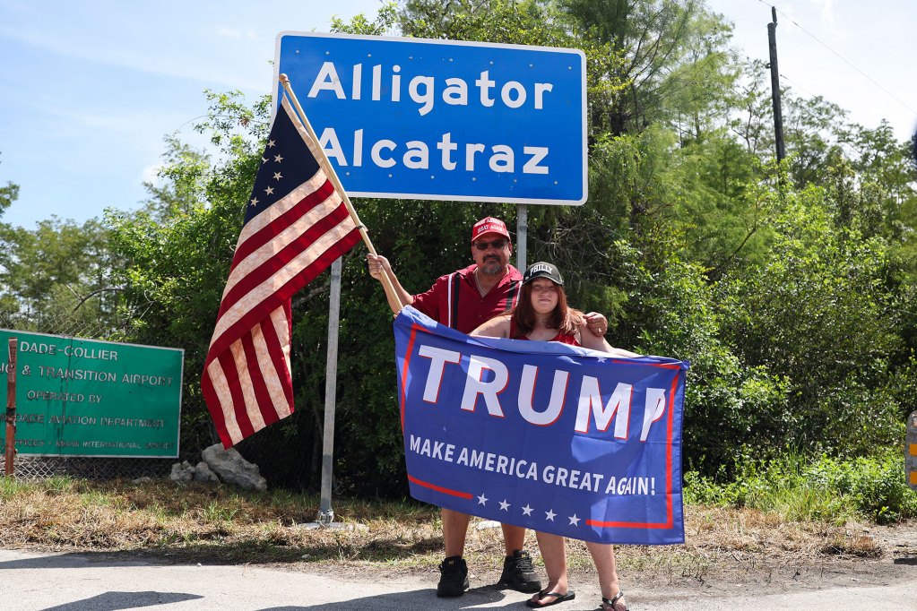 Two people pose with a US Flag and a Trump Flag in front of a sign that reads Alligator Alcatraz