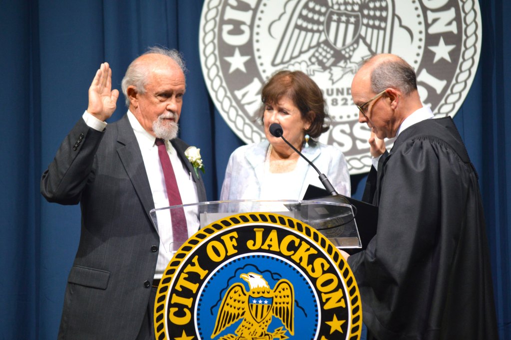 Councilman Ashby Foote wearing a black pinstripe suit with maroon tie stands with his wife while being sworn in by a judge wearing a black robe