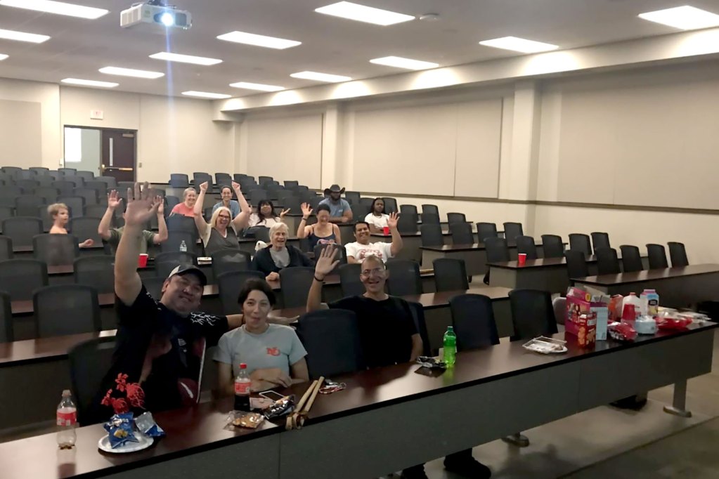 Several people sit in a campus auditorium classroom with snacks and watches something projected on the large screen