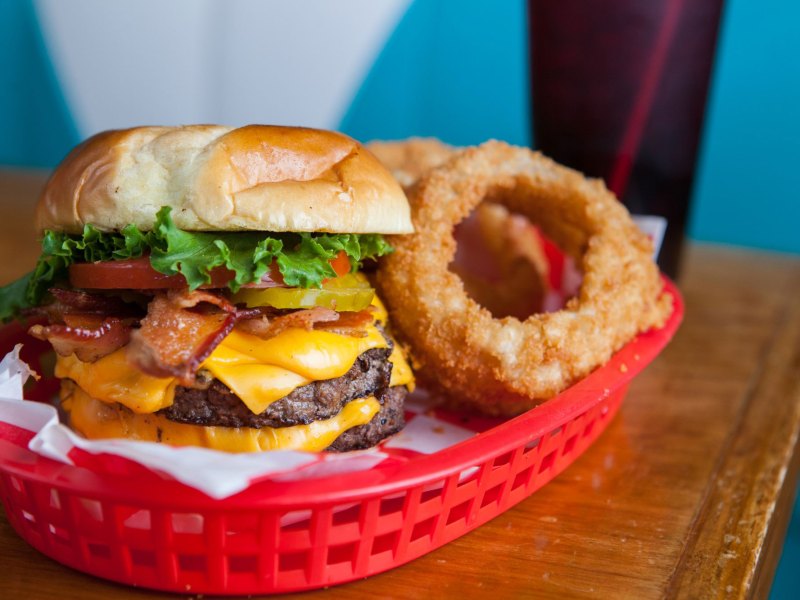 A red basket containing a large hamburger and fried onion rings sits on a wooden table