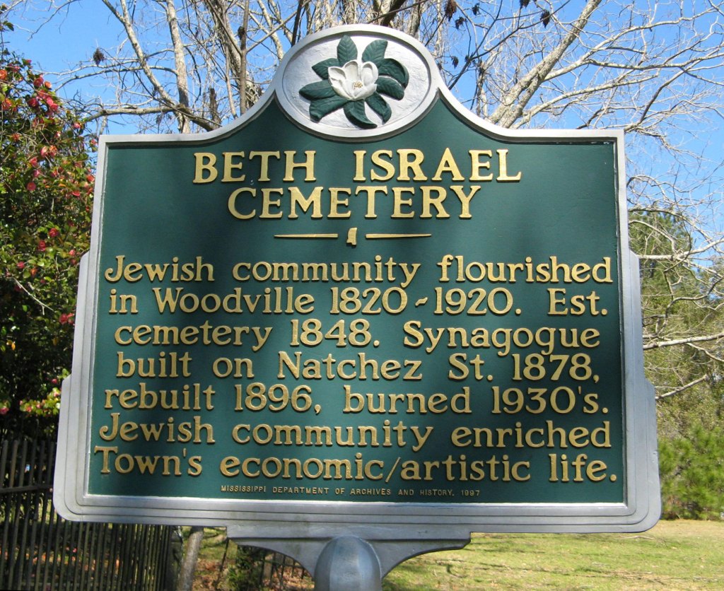 A green history marker outside that reads ‘BETH ISRAEL CEMETERY Jewish community flourished in Woodville 1820 - 1920. Est. cemetery 1848. Synagogue built on Natchez St. 1878, rebuilt 1896, burned 1930's. Jewish community enriched Town's economic/artistic life.‘