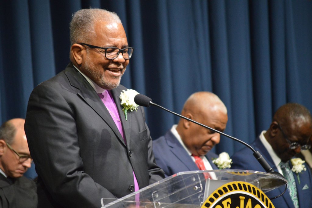 Bishop Ronnie Crudup wearing a black suit with a purple under shirt delivers a speech