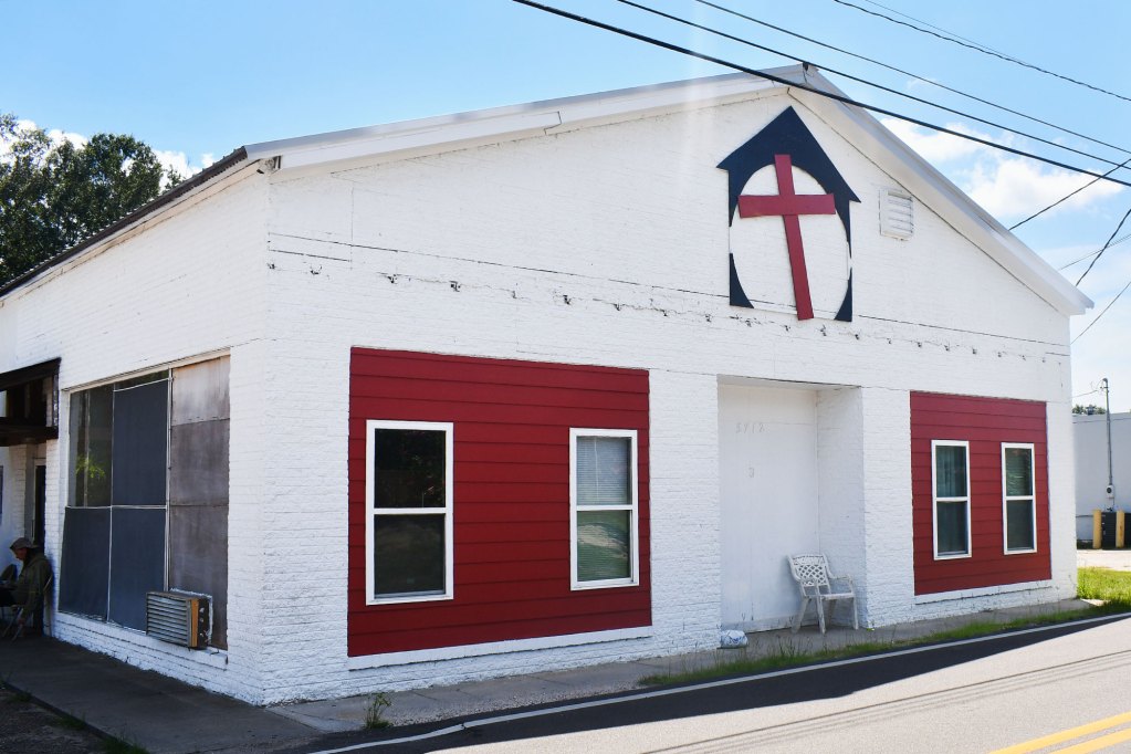 A single-story white brick building with red window panels and a red cross emblem above the front door. The building sits along a roadside under clear skies.