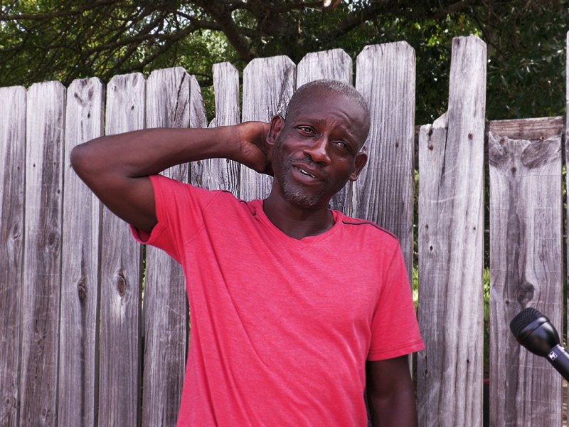A man in a red T-shirt stands in front of a wooden fence with one hand behind his neck, looking toward a microphone held by an interviewer.