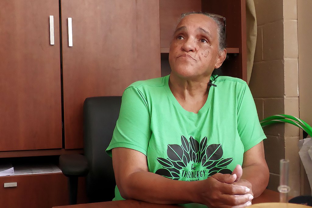 A woman in a green shirt sits at a desk, looking slightly upward. She is seated in an office setting with wooden cabinets behind her.