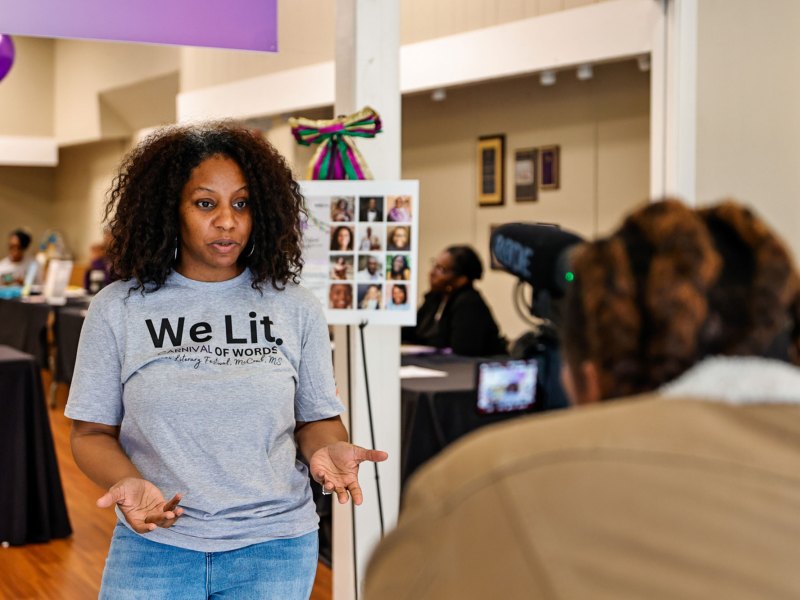 A woman in a shirt that reads 'We Lit' speaks into a camera at an event