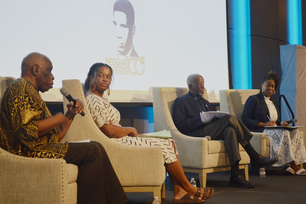 Four people on a stage speaking with a picture of Medgar Evers on a screen behind them