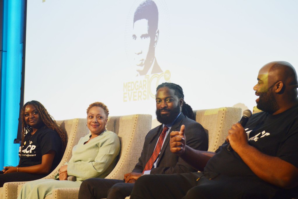 Four people on a stage speaking with a picture of Medgar Evers on a screen behind them