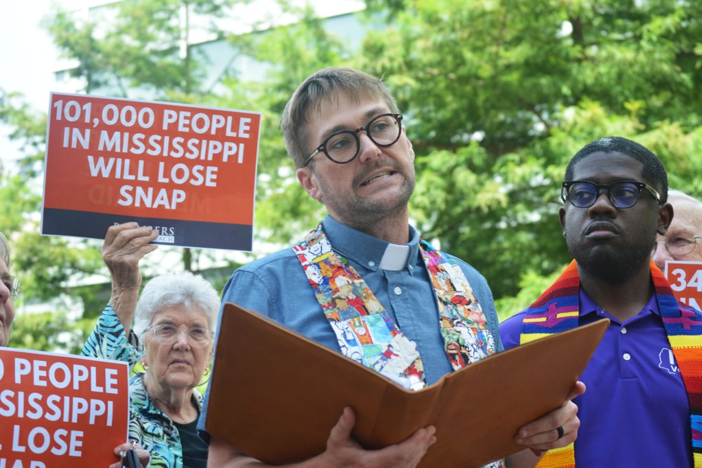 Two faith leaders at a rally, a person holding a sign behind them that reads '101,000 People in Mississippi Will Lose SNAP'