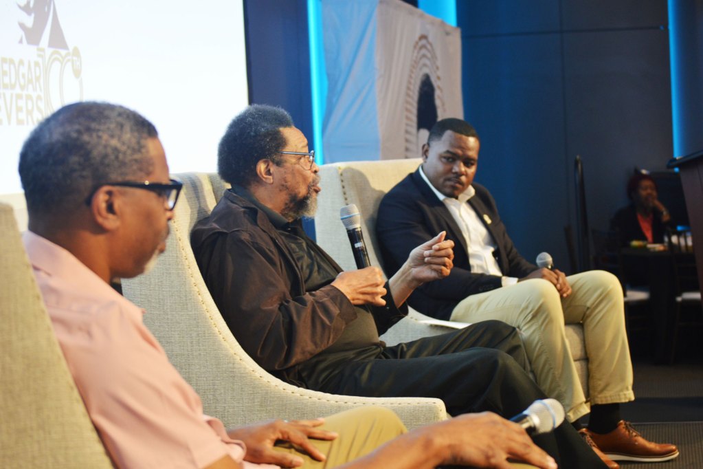 Three men speaking on a stage, one in the middle wearing black and holding a mic